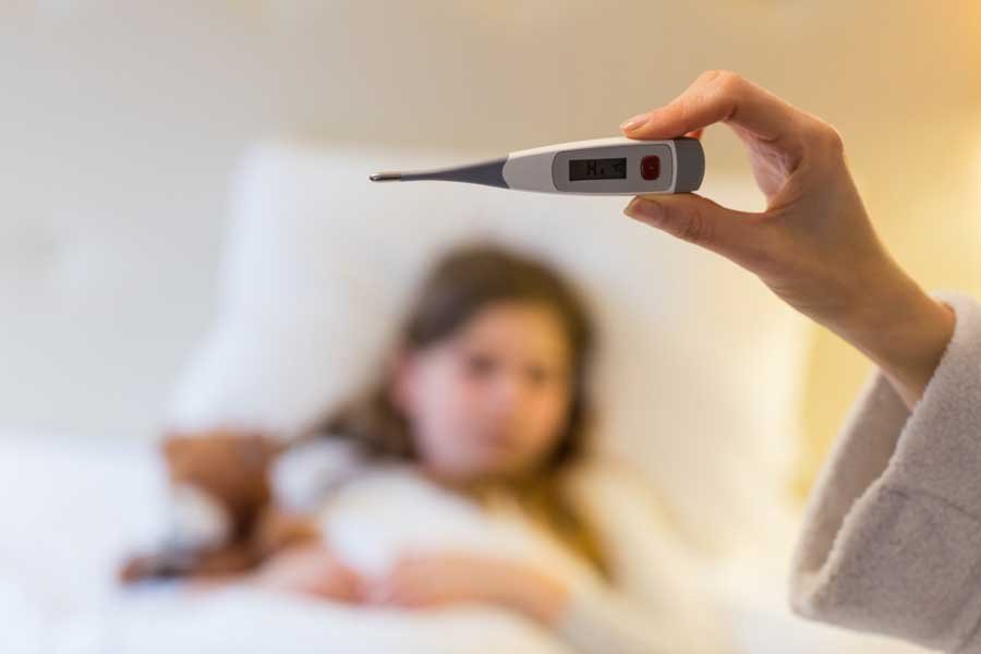 Una niña acostada en la cama enferma con fiebre, mientras una persona le mide la temperatura.