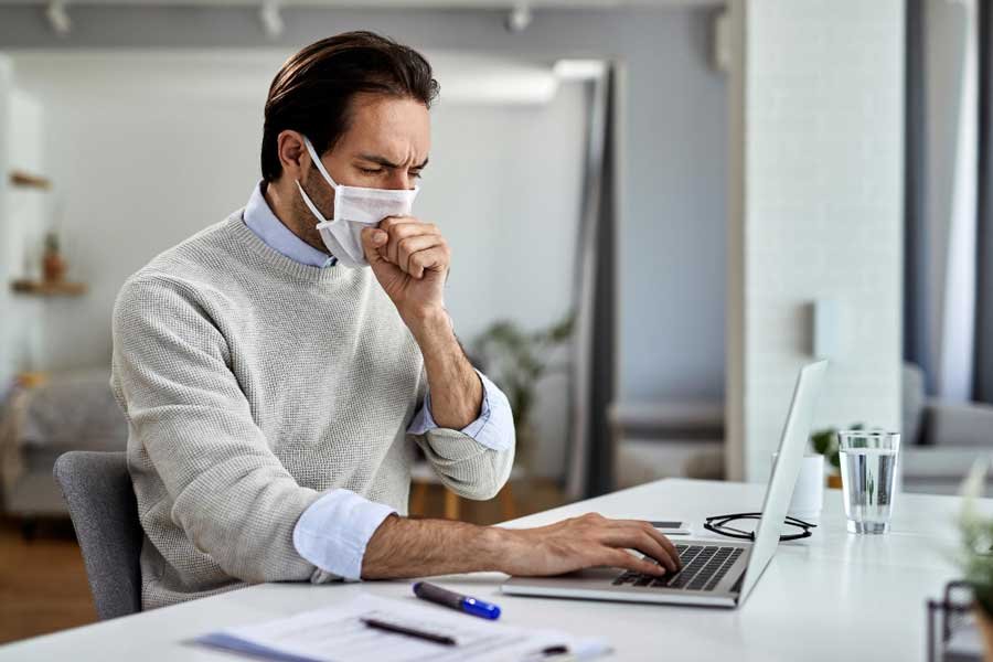 Un hombre sentando frente al computador usando tapabocas, tras presentar síntomas de una infección respiratoria.