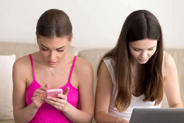 Dos mujeres jóvenes sentadas en un sofá, una con un celular en sus manos y la otra escribiendo en su computador portátil. La postura encorvada de las dos genera dolor de cuello.