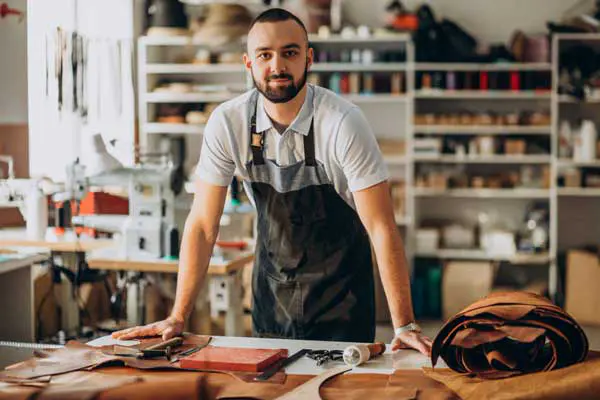Un hombre emprendedor de pie y apoyado en un escritorio, al fondo su emprendimiento de confección de zapatos.
